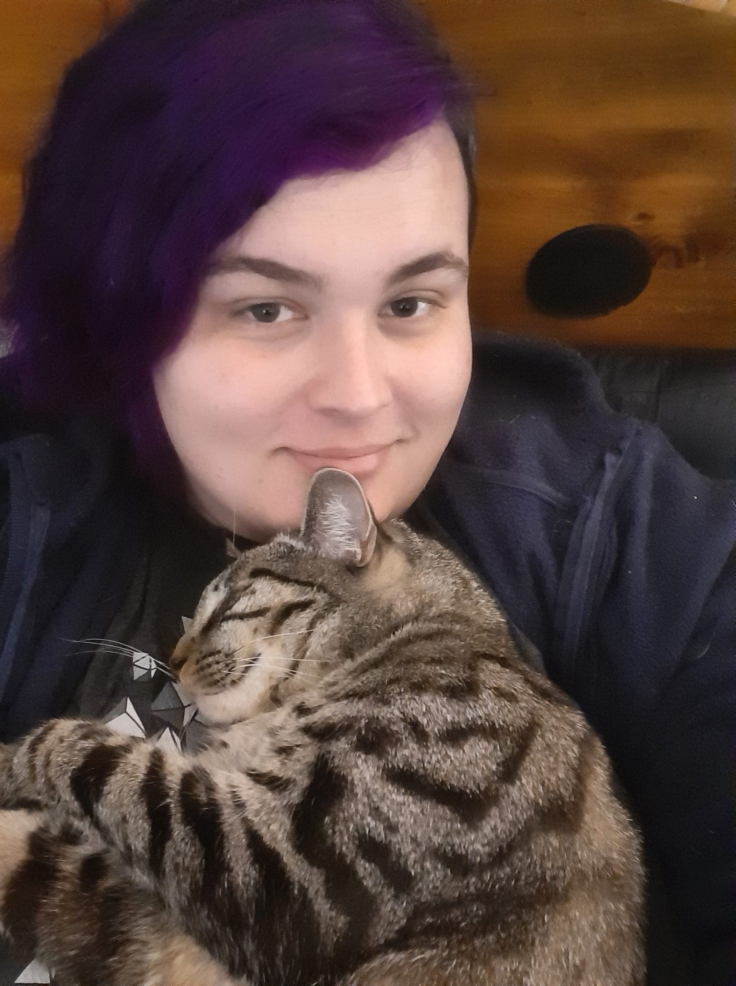 A picture of a white woman with purple hair, smiling, with a tabby cat laying on her chest